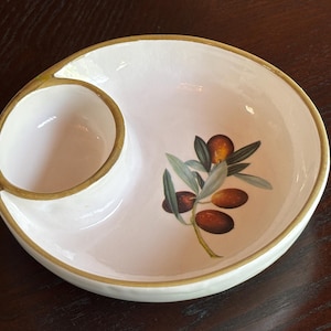 May include: A white ceramic olive dish with a separate compartment. The dish features a painted olive branch design with green leaves and brown olives. The rim and the edge of the small compartment are trimmed with a gold color.