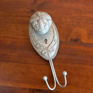 May include: A silver-toned wall hook featuring an oval backplate with a beaded edge and a central keyhole cutout. A round, embossed knob sits at the top of the plate, while two curved metal hooks with rounded ends extend from the bottom section.
