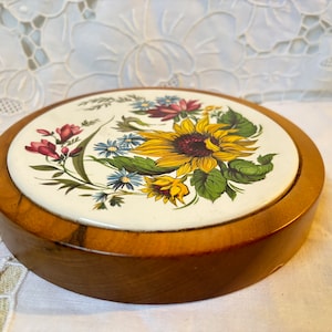 May include: A round wooden box with a floral design on the lid. The lid features a sunflower and other flowers in yellow, red, blue, and green against a white background. The box is made of light brown wood.