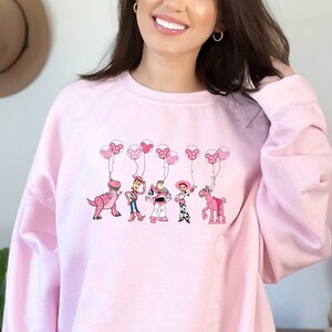 May include: Pink sweatshirt with a graphic of Toy Story characters holding pink balloons. The characters include Woody, Buzz Lightyear, Jessie, and Rex. The text "Shining Colors" is on the bottom left corner of the image.