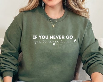 Könnte beinhalten: Eine Frau trägt ein grünes Sweatshirt mit dem Text "IF YOU NEVER GO you'll never know" und einer kleinen Flugzeuggrafik. Sie sitzt auf einem Sofa mit einem grünen Kissen.