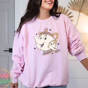 May include: Pink sweatshirt with a cartoon image of Mrs. Potts and Chip from Beauty and the Beast. The image is in a vintage style with gold accents and stars.