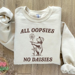 May include: A beige sweatshirt with a brown graphic of a bear holding a daisy. The text "ALL OOPSIESS NO DAISIES" is printed on the sweatshirt.