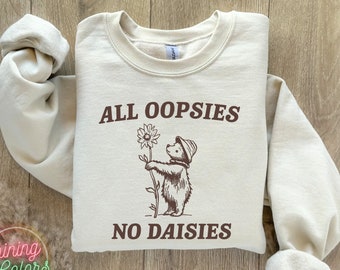 Puede incluir: Una sudadera beige con un gráfico marrón de un oso sosteniendo una margarita. El texto "ALL OOPSIESS NO DAISIES" está impreso en la sudadera.