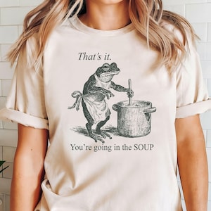 May include: Beige t-shirt featuring a black and white illustration of a frog wearing an apron and stirring a pot. Text above reads "That's it." and below "You're going in the SOUP."