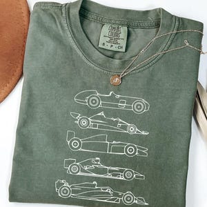 May include: Olive green t-shirt featuring a white line drawing of five race cars. The shirt has a crew neck and short sleeves. A gold necklace with a circular pendant is draped across the shirt.