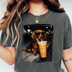 May include: Dark grey t-shirt featuring a dachshund wearing sunglasses and holding an iced coffee. The dog is brown with long fur. The image has a black border and a blurred background of city lights.