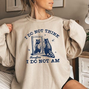 May include: Beige sweatshirt with a raccoon graphic and the text "I DO NOT THINK therefore I DO NOT AM" in navy blue. The raccoon is depicted looking in a mirror. The sweatshirt has long sleeves.