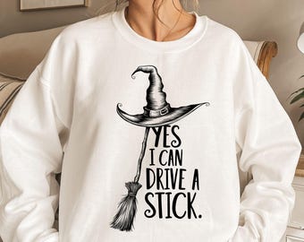 May include: White sweatshirt with a black witch hat and broom graphic. The text "YES I CAN DRIVE A STICK" is printed below the hat and broom. The sweatshirt is a crew neck style.