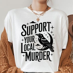 May include: White t-shirt with the text "SUPPORT YOUR LOCAL MURDER" in black, featuring a black raven perched on a crescent moon. The design includes stars and a cloud. The shirt is part of a clothing collection.