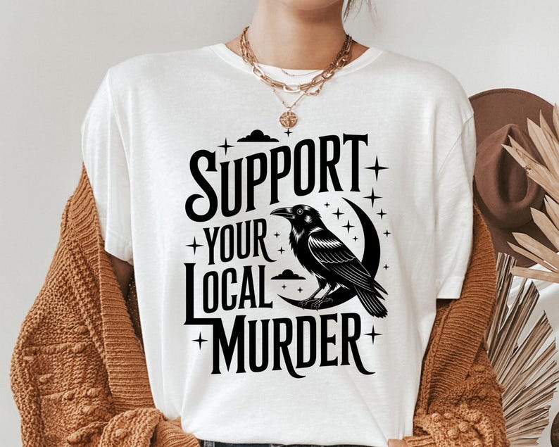 May include: White t-shirt with the text "SUPPORT YOUR LOCAL MURDER" in black, featuring a black raven perched on a crescent moon. The design includes stars and a cloud. The shirt is part of a clothing collection.