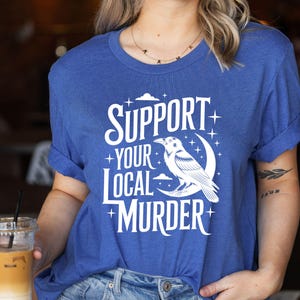 May include: A blue t-shirt with the text "SUPPORT YOUR LOCAL MURDER" in white, along with a graphic of a raven perched on a crescent moon. The shirt has rolled-up sleeves.