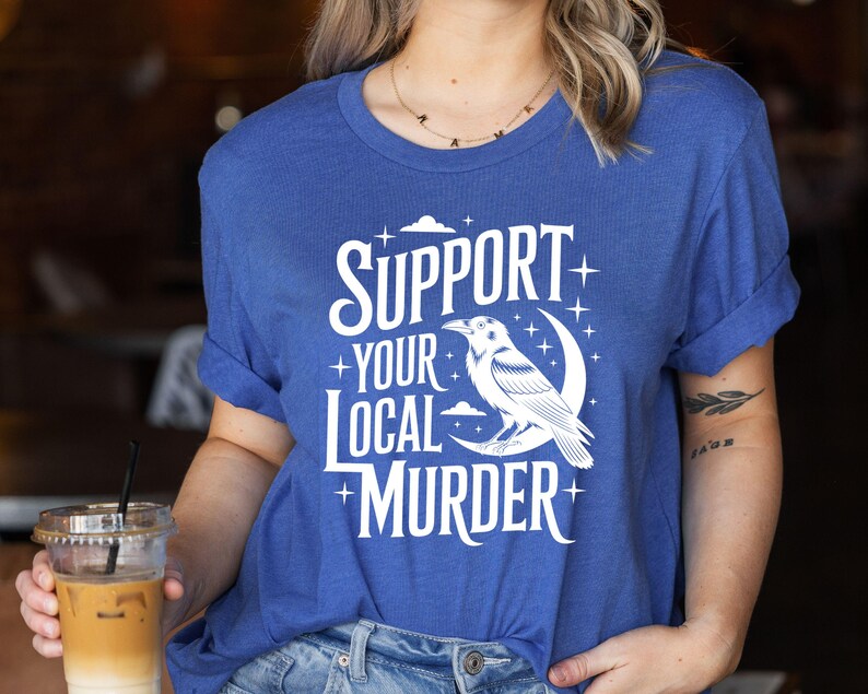 May include: A blue t-shirt with the text "SUPPORT YOUR LOCAL MURDER" in white, along with a graphic of a raven perched on a crescent moon. The shirt has rolled-up sleeves.