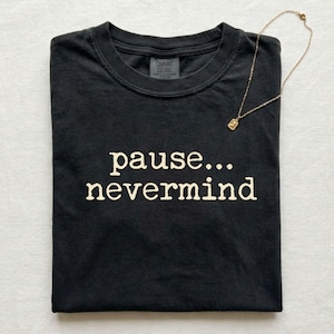 May include: A black short-sleeved t-shirt featuring the white text "pause... nevermind" printed on the chest. A thin gold-toned chain necklace with a small rectangular pendant rests on the upper right side of the folded garment.