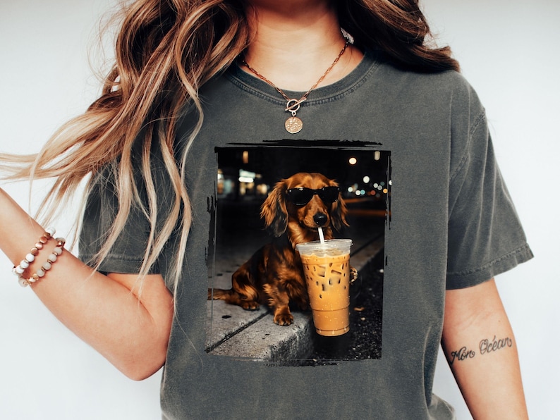 May include: Dark grey t-shirt featuring a dachshund wearing sunglasses and holding an iced coffee. The dog is brown with long fur. The image has a black border and a blurred background of city lights.