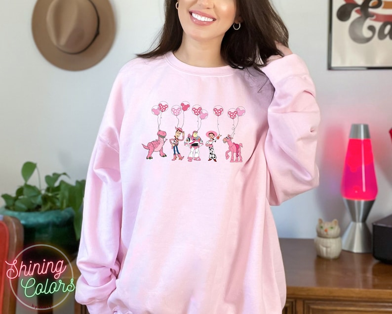 May include: Pink sweatshirt with a graphic of Toy Story characters holding pink balloons. The characters include Woody, Buzz Lightyear, Jessie, and Rex. The text "Shining Colors" is on the bottom left corner of the image.