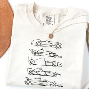 K&ouml;nnte beinhalten: Cremefarbenes T-Shirt mit einer schwarzen Strichzeichnung von f&uuml;nf Oldtimer-Rennwagen. Das Shirt hat einen Rundhalsausschnitt und kurze &Auml;rmel. Eine goldene Halskette mit Anh&auml;nger liegt auf dem Shirt. Das Etikett lautet "COMFORT COLORS".