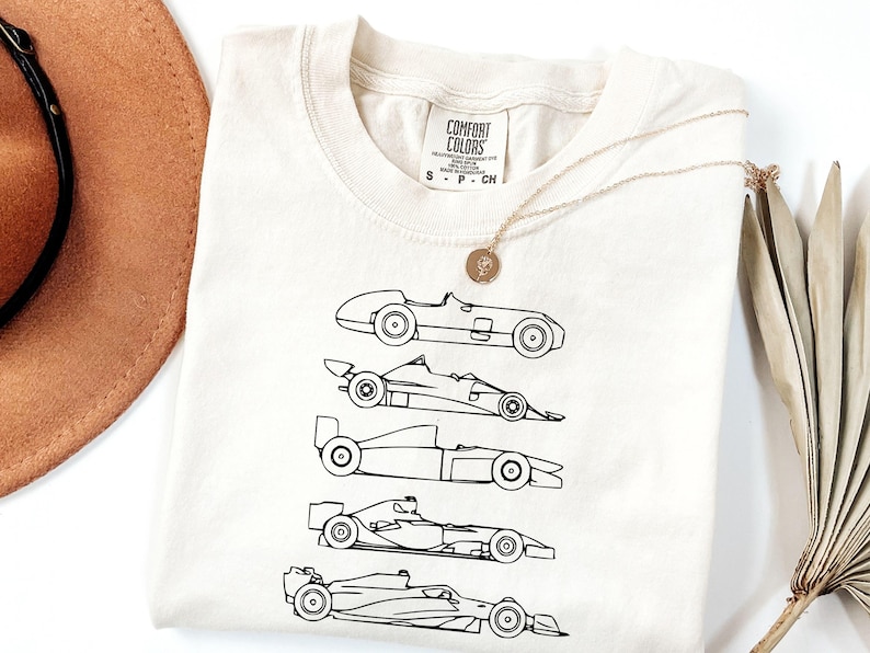 K&ouml;nnte beinhalten: Cremefarbenes T-Shirt mit einer schwarzen Strichzeichnung von f&uuml;nf Oldtimer-Rennwagen. Das Shirt hat einen Rundhalsausschnitt und kurze &Auml;rmel. Eine goldene Halskette mit Anh&auml;nger liegt auf dem Shirt. Das Etikett lautet "COMFORT COLORS".