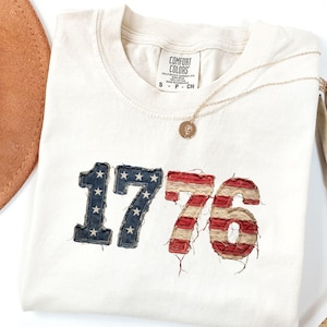 Puede incluir: Camiseta color crema con el número "1776" en un diseño patriótico. El "17" es azul con estrellas blancas, y el "76" es de rayas rojas y blancas. Un collar dorado con un colgante está sobre la camiseta.