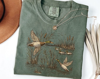 May include: Sage green Comfort Colors t-shirt featuring a detailed illustration of ducks in a marsh setting. The design includes flying ducks, reeds, and a pond with ducks swimming. A gold necklace and a brown hat are also visible.