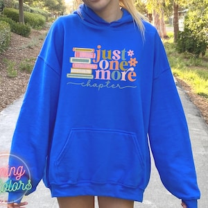 May include: A royal blue hoodie featuring a stack of colourful books and the text "just one more chapter" in a whimsical font.