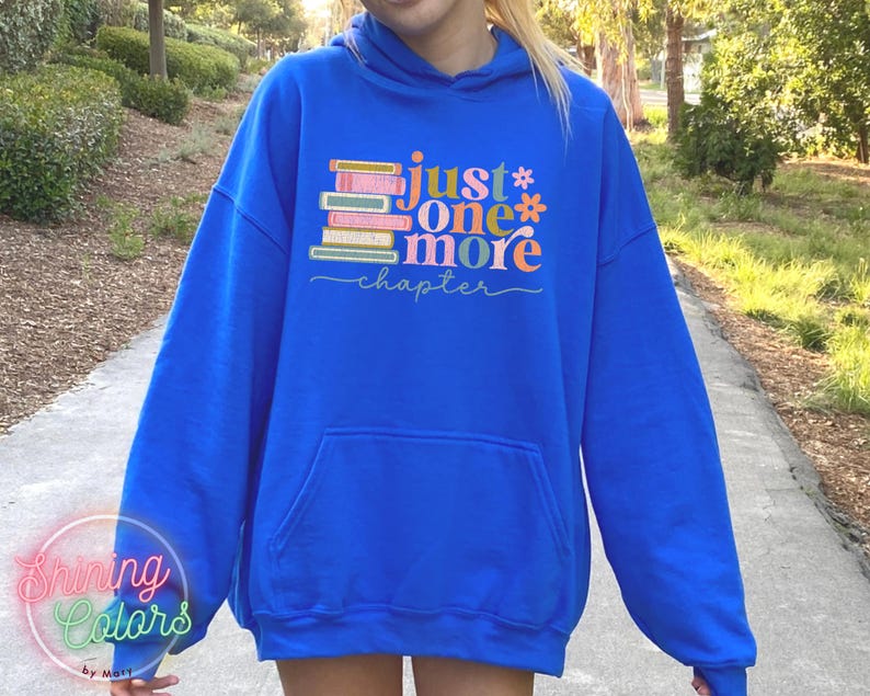 May include: A royal blue hoodie featuring a stack of colourful books and the text "just one more chapter" in a whimsical font.