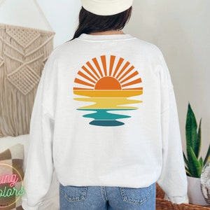 May include: White sweatshirt with a retro sunset graphic on the back. The graphic features a yellow sun with orange rays and a blue and green reflection in the water.