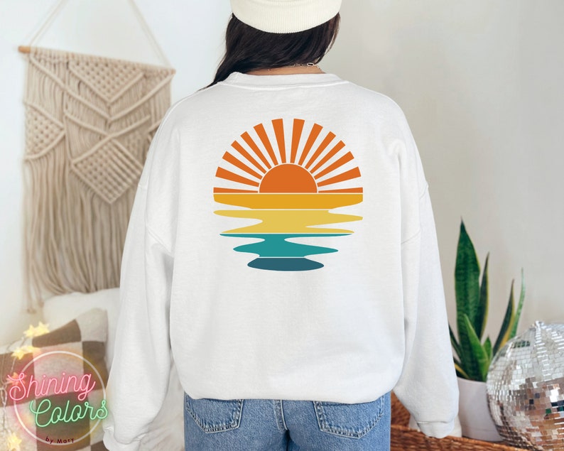 May include: White sweatshirt with a retro sunset graphic on the back. The graphic features a yellow sun with orange rays and a blue and green reflection in the water.