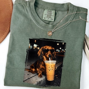 May include: Olive green Comfort Colors t-shirt with a graphic of a dachshund wearing sunglasses and drinking an iced coffee. A gold necklace and a small tag are on the shirt. A brown hat and a dried palm leaf are in the background.