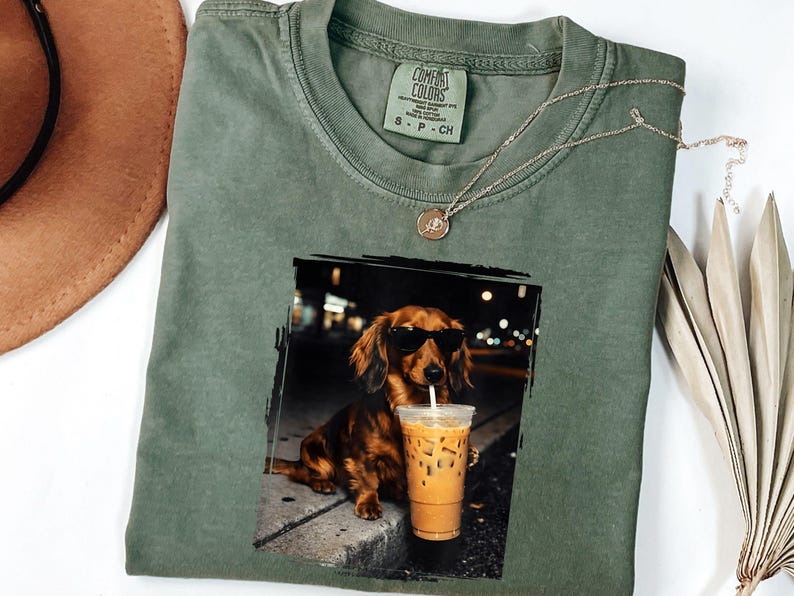 May include: Olive green Comfort Colors t-shirt with a graphic of a dachshund wearing sunglasses and drinking an iced coffee. A gold necklace and a small tag are on the shirt. A brown hat and a dried palm leaf are in the background.