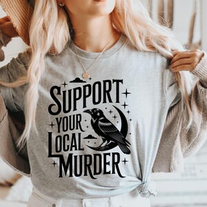 May include: A light gray t-shirt with the text "SUPPORT YOUR LOCAL MURDER" in black. A black raven is perched on a crescent moon. The design includes stars and clouds.