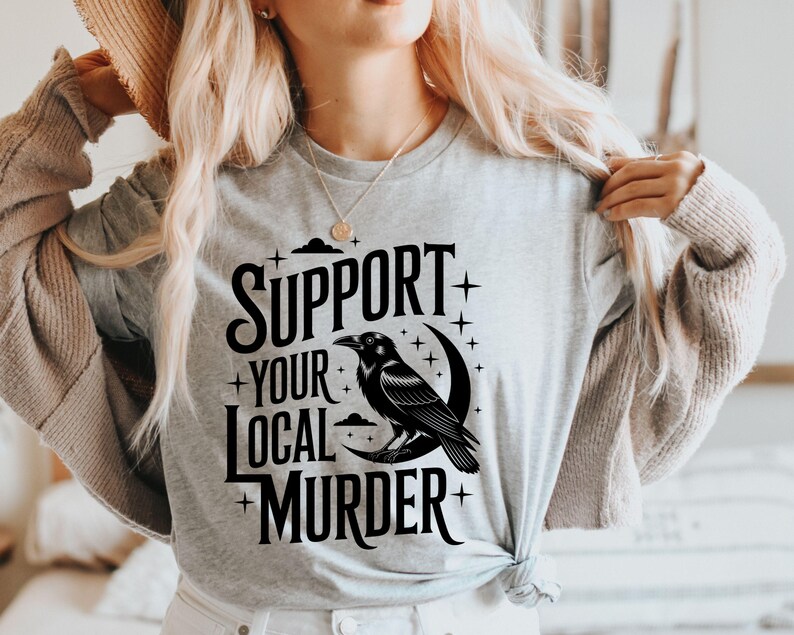 May include: A light gray t-shirt with the text "SUPPORT YOUR LOCAL MURDER" in black. A black raven is perched on a crescent moon. The design includes stars and clouds.