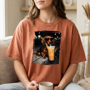 May include: An orange t-shirt featuring a graphic of a dachshund wearing sunglasses and holding an iced coffee. The dog is sitting on a curb in front of a dark background. The shirt is a solid color.