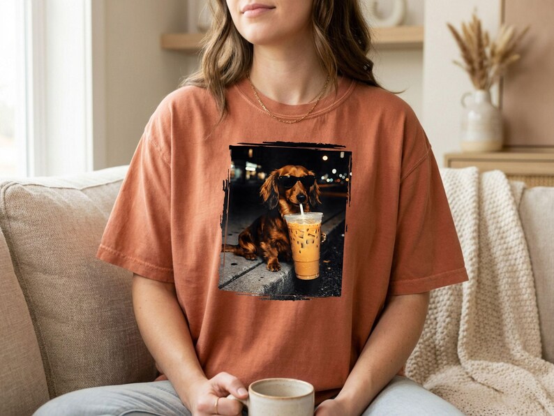 May include: An orange t-shirt featuring a graphic of a dachshund wearing sunglasses and holding an iced coffee. The dog is sitting on a curb in front of a dark background. The shirt is a solid color.