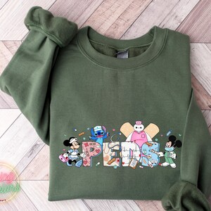 May include: Green sweatshirt with a graphic print of Disney characters dressed as nurses. The characters include Stitch, Mickey Mouse, Baymax, and a nurse Minnie Mouse. The text "NURSES" is spelled out with medical symbols and bandages.