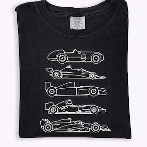 K&ouml;nnte beinhalten: Schwarzes T-Shirt mit einer wei&szlig;en Umrisszeichnung von f&uuml;nf Rennwagen. Das Comfort Colors-Etikett ist oben sichtbar. Die Autos sind vertikal angeordnet und zeigen verschiedene Designs aus verschiedenen Rennepochen. Das T-Shirt ist gefaltet.