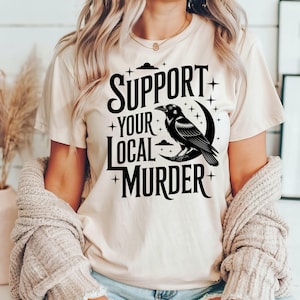May include: Beige t-shirt with the text "SUPPORT YOUR LOCAL MURDER" in black, featuring a raven perched on a crescent moon. The design includes stars and cloud accents. The shirt is a casual, everyday wear item.