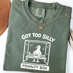May include: Olive green t-shirt with the text "GOT TOO SILLY PENALTY BOX" and a graphic of a duck in a penalty box. A gold necklace with a pendant and a brown hat are also visible.