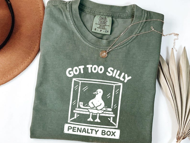 May include: Olive green t-shirt with the text "GOT TOO SILLY PENALTY BOX" and a graphic of a duck in a penalty box. A gold necklace with a pendant and a brown hat are also visible.