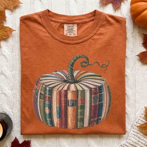 May include: A burnt orange short-sleeve t-shirt featuring a graphic of a pumpkin constructed from a circular arrangement of vintage books with a green stem. The shirt has a crew neckline and a Comfort Colors brand label printed on the inside collar.