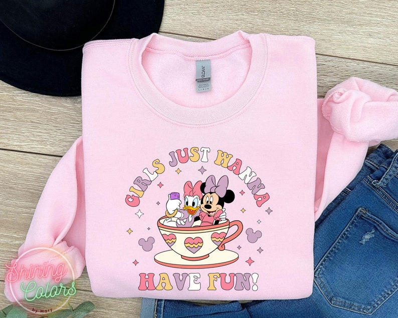 Puede incluir: Sudadera rosa con un gr&aacute;fico de Minnie Mouse y Daisy Duck en una taza de t&eacute;. El texto "Girls Just Wanna Have Fun!" est&aacute; impreso debajo de la imagen.