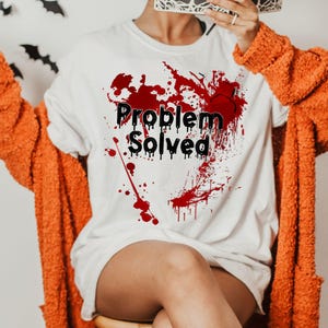 May include: White t-shirt with the text "Problem Solved" in black, with red splatter design. The person is wearing an orange cardigan. Black bat decorations are in the background.