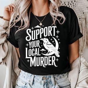 May include: Black t-shirt with the text "SUPPORT YOUR LOCAL MURDER" in white, featuring a white raven perched on a crescent moon. The design includes stars and cloud details, creating a gothic aesthetic.