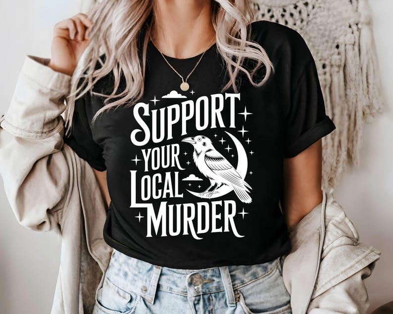 May include: Black t-shirt with the text "SUPPORT YOUR LOCAL MURDER" in white, featuring a white raven perched on a crescent moon. The design includes stars and cloud details, creating a gothic aesthetic.