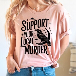 May include: A light pink t-shirt with the text "SUPPORT YOUR LOCAL MURDER" in black, along with a black raven perched on a crescent moon. The design includes stars and clouds.