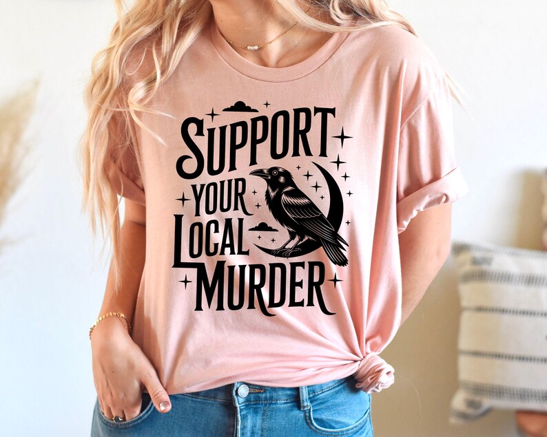 May include: A light pink t-shirt with the text "SUPPORT YOUR LOCAL MURDER" in black, along with a black raven perched on a crescent moon. The design includes stars and clouds.