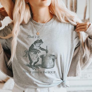 May include: Light gray long-sleeve shirt featuring a frog chef stirring a pot of soup. The text "That's it. You're going in the SOUP" is printed above and below the frog illustration. A humorous design.