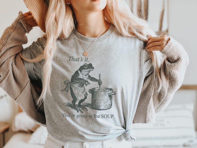 May include: Light gray long-sleeve shirt featuring a frog chef stirring a pot of soup. The text "That's it. You're going in the SOUP" is printed above and below the frog illustration. A humorous design.