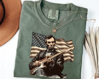 May include: Green t-shirt featuring an illustration of Abraham Lincoln playing an electric guitar in front of an American flag. The shirt has a crew neck and short sleeves. A gold necklace is draped across the shirt.
