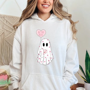 May include: White hoodie with a pink ghost holding a pink heart balloon that says "Be My Boo". The ghost is covered in small pink hearts.